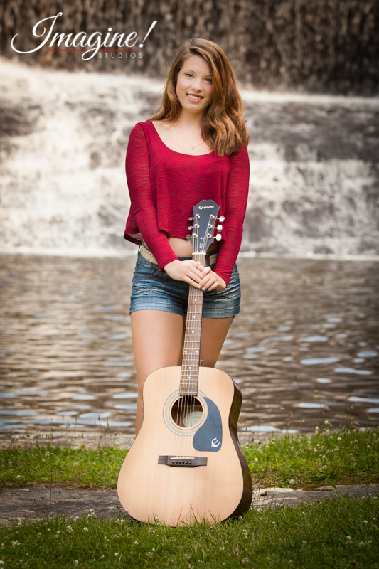 Bethany stands with her guitar by a pond during her senior model outdoor photo session at Highland Lake Inn in Flat Rock, NC